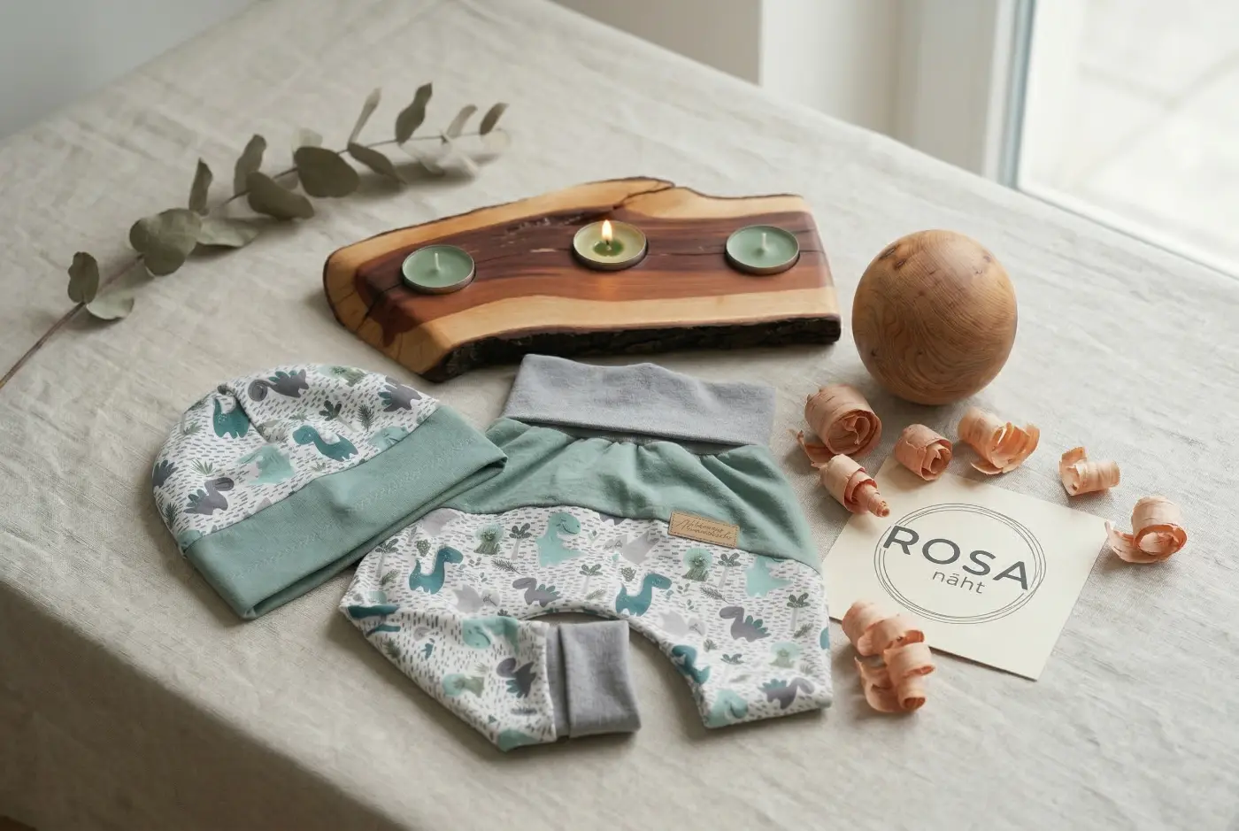 Ein handgemachtes Baby-Outfit Set bestehend aus einer Pumphose und passender Beanie-Mütze mit niedlichem Dinosaurier-Muster in Mintgrün und Grau. Das Set liegt auf einer Leinendecke neben einem rustikalen Teelichthalter aus Naturholz, einer gedrechselten Holzkugel und dekorativen Holzspänen.