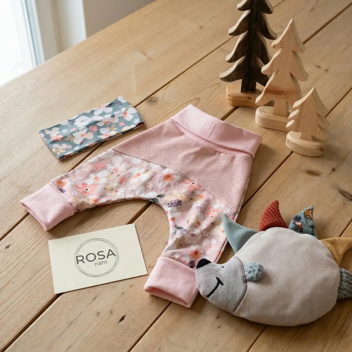 Handgemachtes Baby-Set von ROSA näht, bestehend aus einer rosa Pumphose mit Blumenmuster, einem passenden Stirnband und einem Igel-Schmusetuch auf einem Holztisch mit Deko-Tannenbäumen.