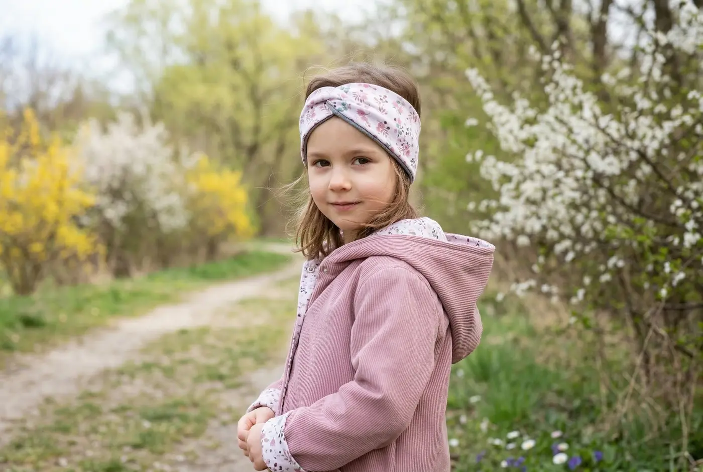 Ein kleines Mädchen im Freien trägt eine handgenähte altrosa Cordjacke mit Kapuze und ein florales Stirnband vor blühenden Frühlingssträuchern.
