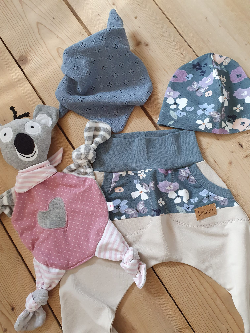 Ein handgefertigtes Babyset von ROSA näht auf einem Holzboden. Es besteht aus einer mitwachsenden Pumphose und einer passenden Mütze in Blau- und Beigetönen mit floralem Schmetterlingsmuster, ergänzt durch ein kleines blaues Halstuch und ein süßes Koala-Kuscheltier mit appliziertem Herz auf dem pinken Bauch.