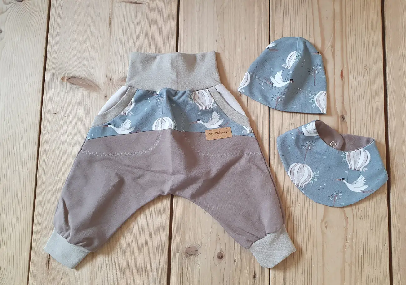 Ein handgenähtes Baby-Set von ROSA näht, flach liegend auf einem hellen Holzboden. Es besteht aus einer mitwachsenden Pumphose und einer passenden Beanie-Mütze. Der Stoff der Mütze und des oberen Hosenteils zeigt eine illustrierte Weltkarte mit verschiedenen Tieren auf hellblauem Grund. Die Hose wird ergänzt durch einen cremefarbenen Stoff im unteren Bereich, taubenblaue Bündchen, kleine Taschen und ein braunes Kunstleder-Label mit der Aufschrift "Kleiner LAUSER".