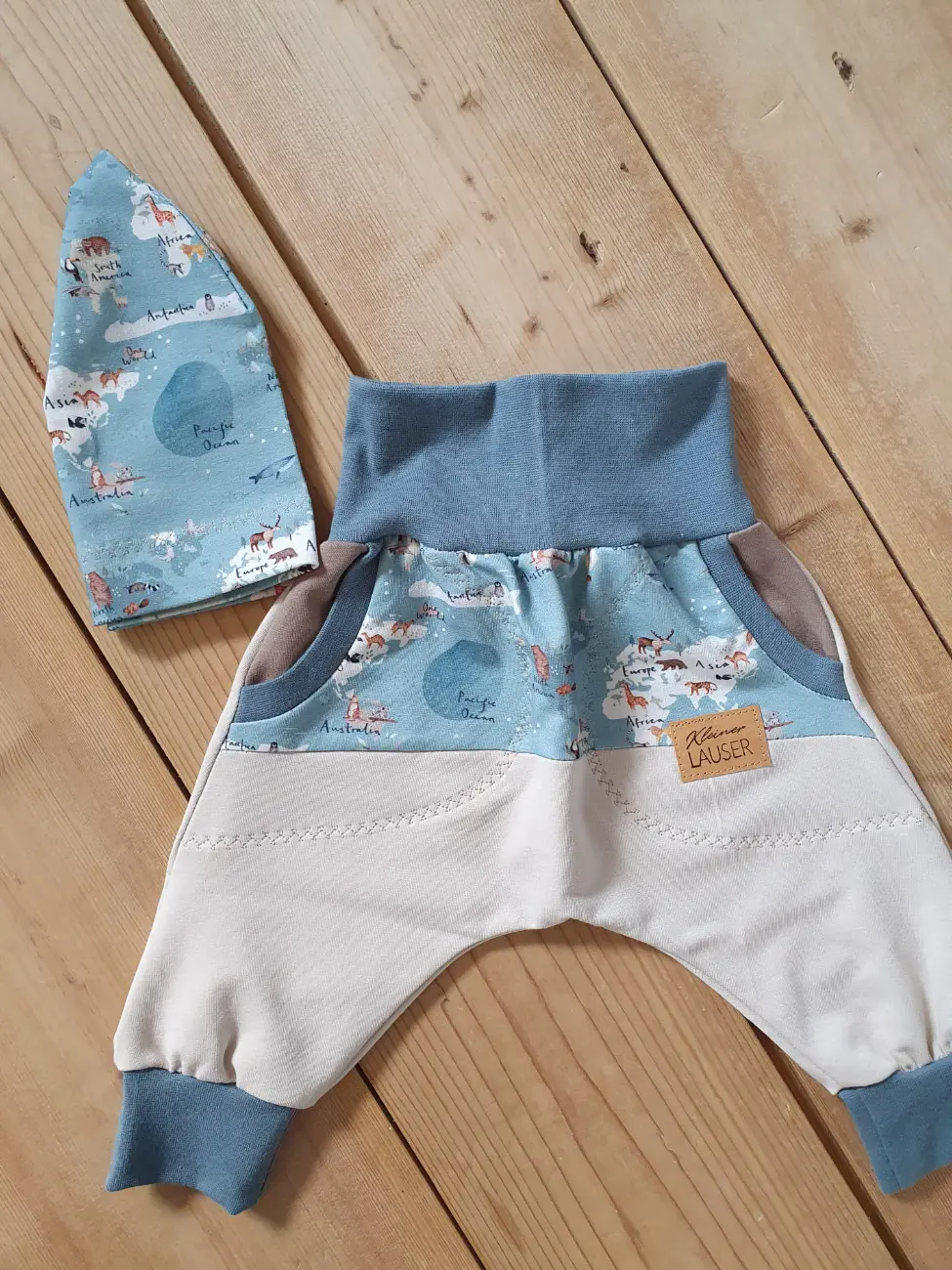 Ein handgenähtes Baby-Set von ROSA näht, flach liegend auf einem hellen Holzboden. Es besteht aus einer mitwachsenden Pumphose und einer passenden Beanie-Mütze. Der Stoff der Mütze und des oberen Hosenteils zeigt eine illustrierte Weltkarte mit verschiedenen Tieren auf hellblauem Grund. Die Hose wird ergänzt durch einen cremefarbenen Stoff im unteren Bereich, taubenblaue Bündchen, kleine Taschen und ein braunes Kunstleder-Label mit der Aufschrift "Kleiner LAUSER".