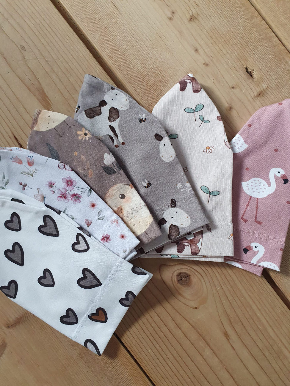 Mehrere handgenähte Babymützen von ROSA näht, aufgefächert auf einem hellen Holzboden liegend. Die weichen Jerseystoffe zeigen liebevolle Motive wie Herzen, Blumen, kleine Kühe und rosafarbene Flamingos.