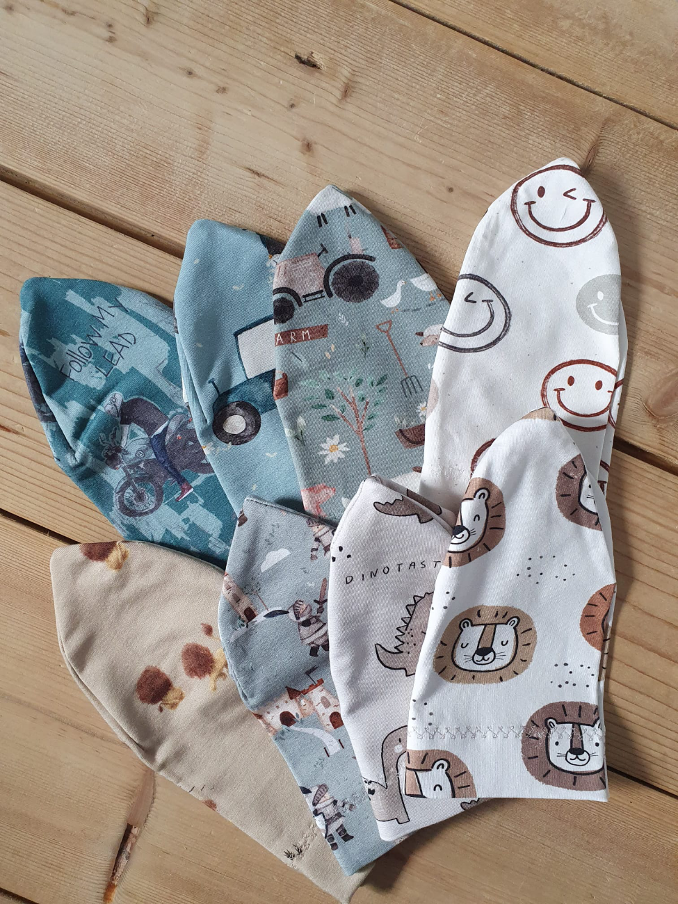 Eine bunte Auswahl an handgenähten Mützen und Beanies von ROSA näht, fächerförmig auf einem Holzboden drapiert. Die Mützen zeigen verschiedene verspielte Muster wie Traktoren, Dinosaurier, Löwen, Smileys und Motorräder.