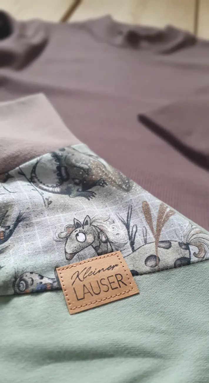 Ein handgefertigter langärmeliger Kinderpullover mit vintage Blumenmuster und dunkelgrünen Bündchen sowie eine cremefarbene Pumphose von ROSA näht, getragen von einem Mädchen auf einer grünen Wiese vor einem Tipi.