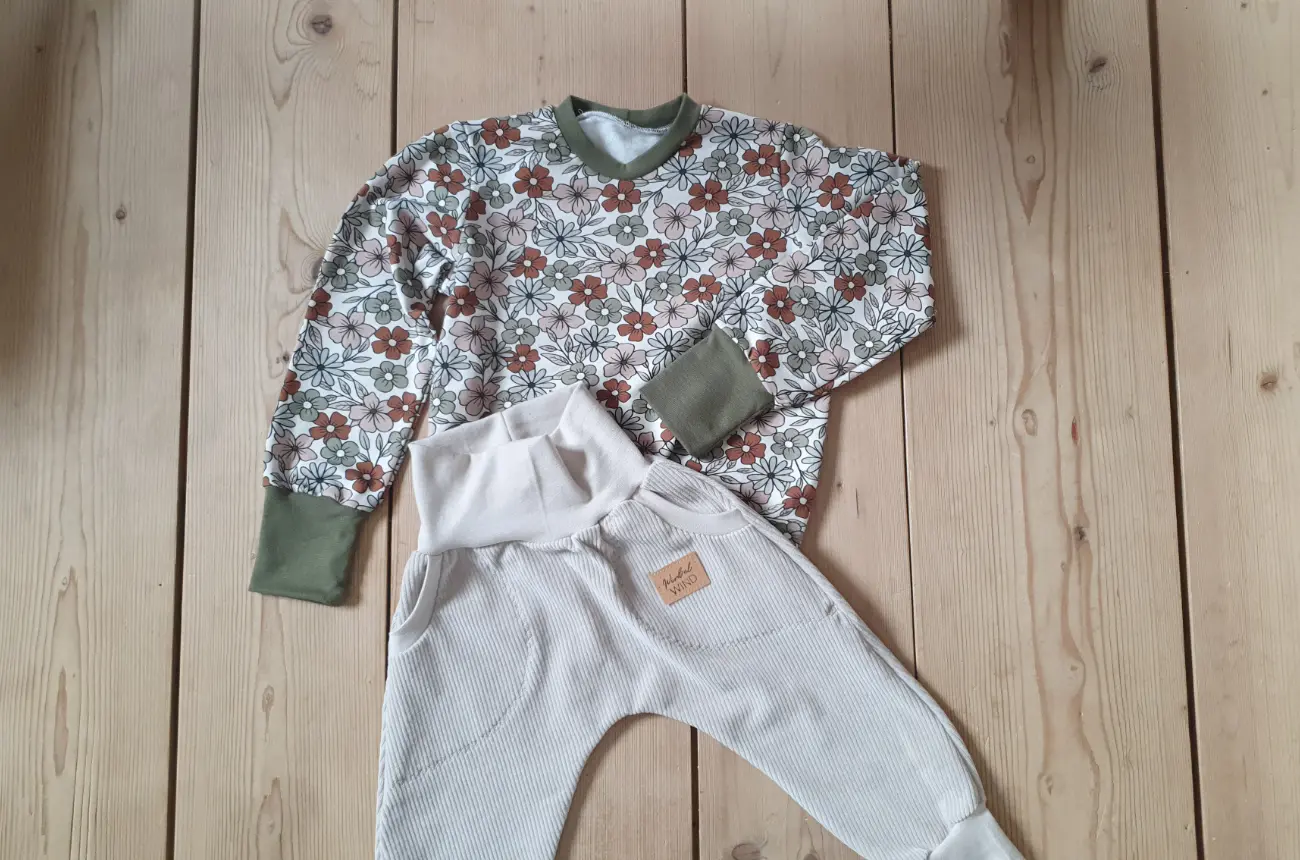 Ein handgenähtes Kinder-Outfit von ROSA näht auf einem hellen Holzboden. Zu sehen ist ein Langarmshirt mit einem Retro-Blumenmuster in Erdtönen und olivgrünen Bündchen, wunderschön kombiniert mit einer bequemen beigen Pumphose aus Cord mit aufgesetzten Taschen.