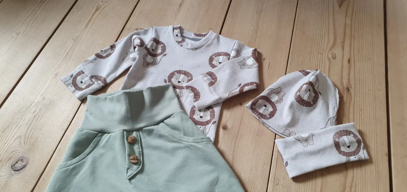 Ein handgenähtes Kinderset von ROSA näht auf einem Holzboden. Es besteht aus einer bequemen mintgrünen Hose mit dekorativen Holzknöpfen, einem Langarmshirt, einer Mütze und einem Loop aus hellem Stoff mit niedlichem Löwen-Muster.