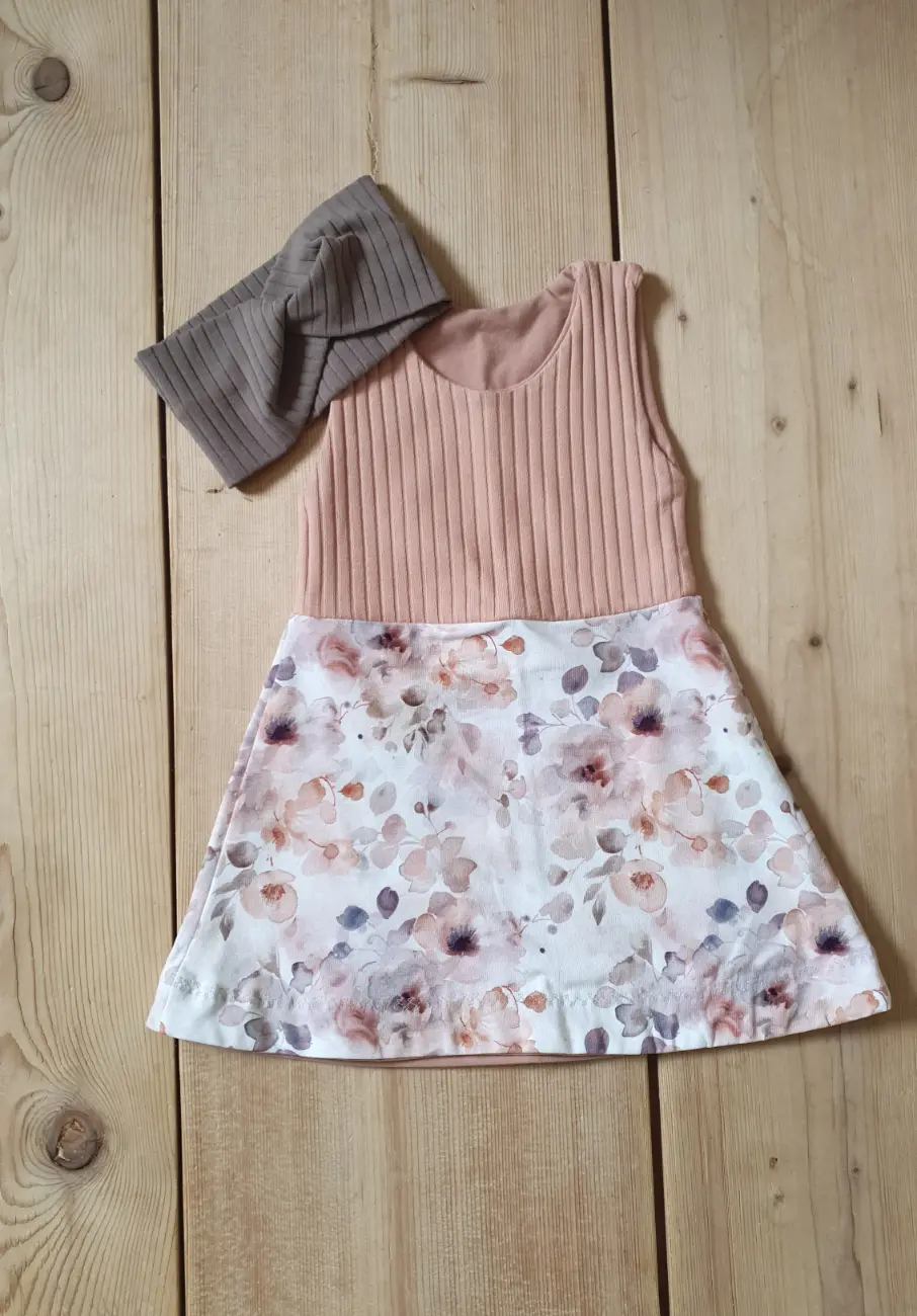 Ein Kinderkleid von ROSA näht mit einem Oberteil aus altrosa Rippjersey und einem angenähten Rockteil mit floralem Muster. Daneben liegt ein farblich passendes Knotenstirnband aus braunem Rippjersey auf dem Holzboden.