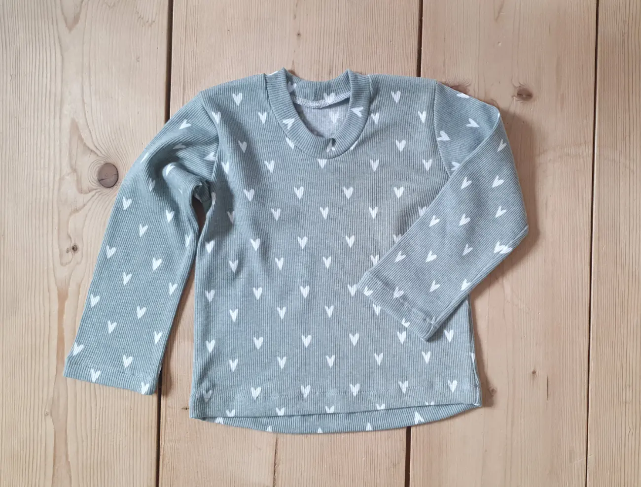 Ein handgefertigter langärmeliger Kinderpullover mit vintage Blumenmuster und dunkelgrünen Bündchen sowie eine cremefarbene Pumphose von ROSA näht, getragen von einem Mädchen auf einer grünen Wiese vor einem Tipi.