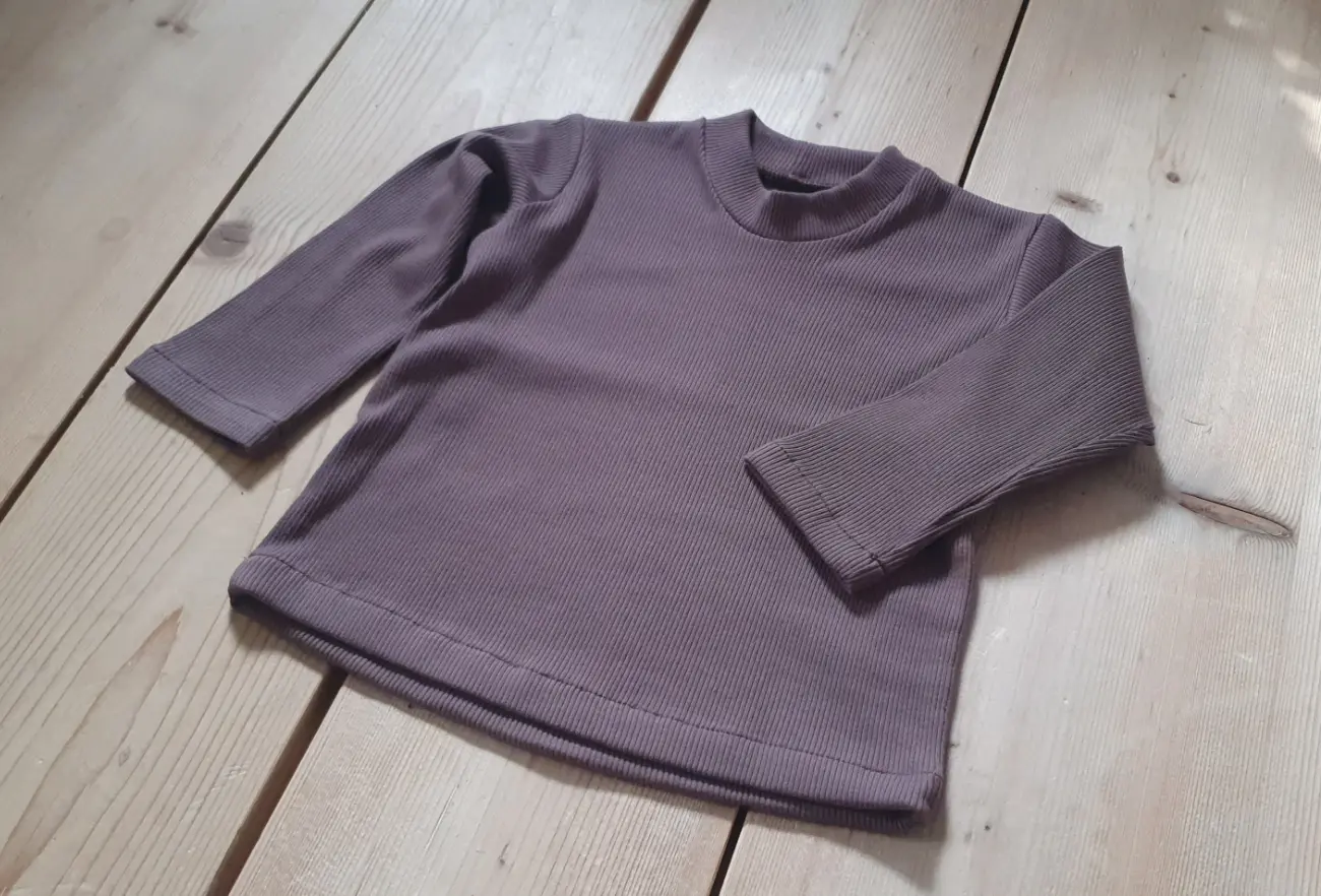 Ein schlichtes, langärmeliges Kindershirt aus dehnbarem, violettem Rippjersey von ROSA näht, flach liegend auf einem hellen Holzboden.