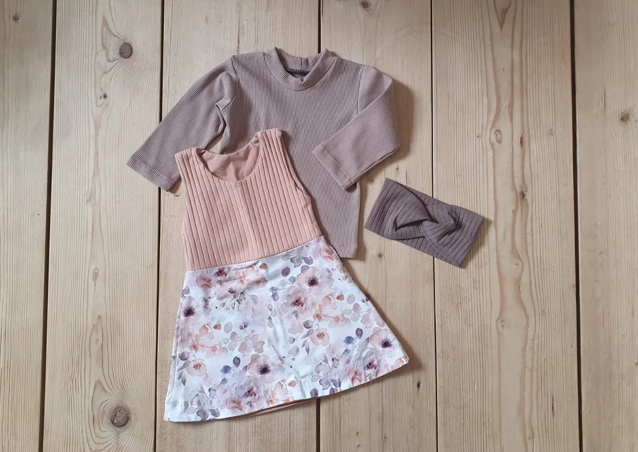 Ein handgefertigter langärmeliger Kinderpullover mit vintage Blumenmuster und dunkelgrünen Bündchen sowie eine cremefarbene Pumphose von ROSA näht, getragen von einem Mädchen auf einer grünen Wiese vor einem Tipi.