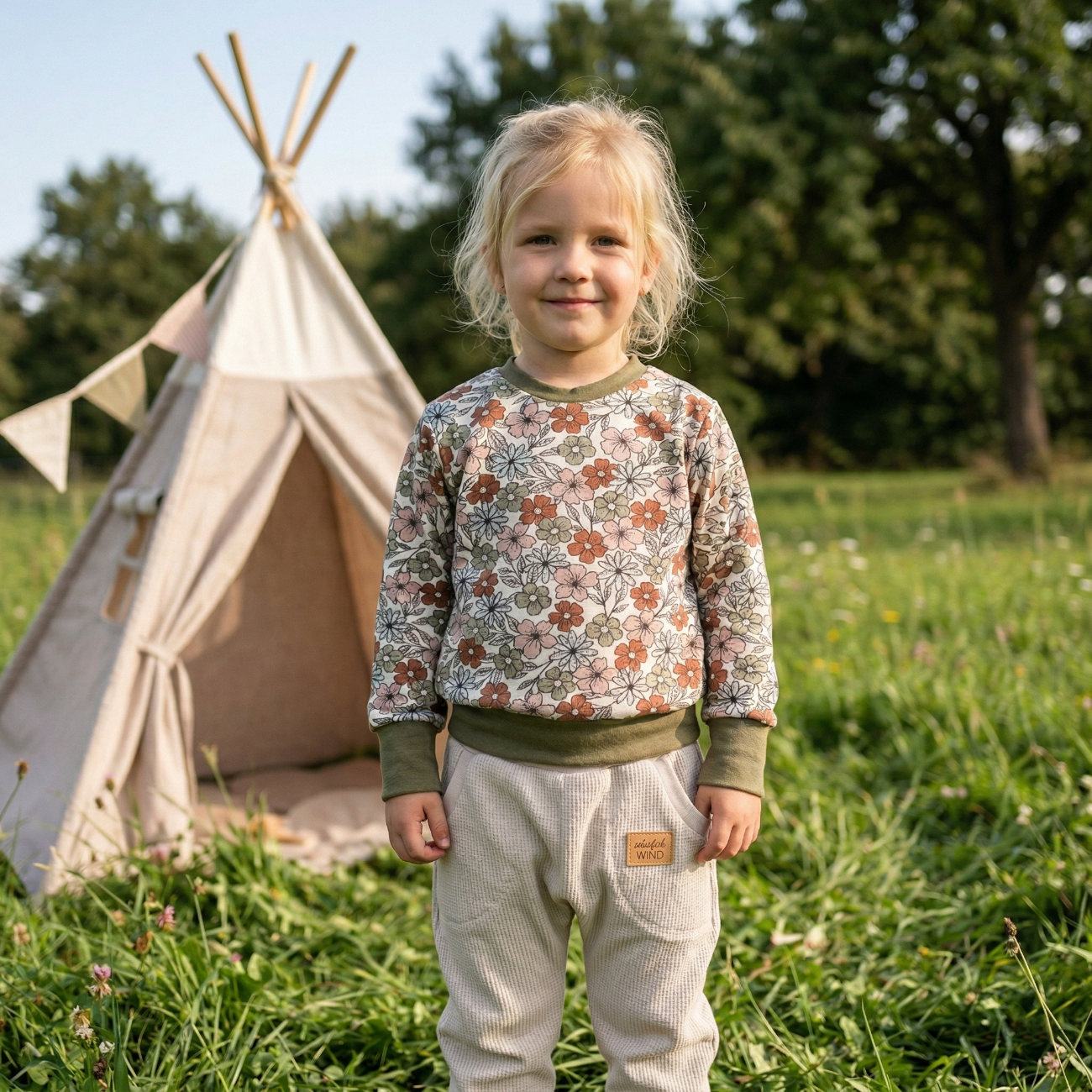 Ein handgefertigter langärmeliger Kinderpullover mit vintage Blumenmuster und dunkelgrünen Bündchen sowie eine cremefarbene Pumphose von ROSA näht, getragen von einem Mädchen auf einer grünen Wiese vor einem Tipi.