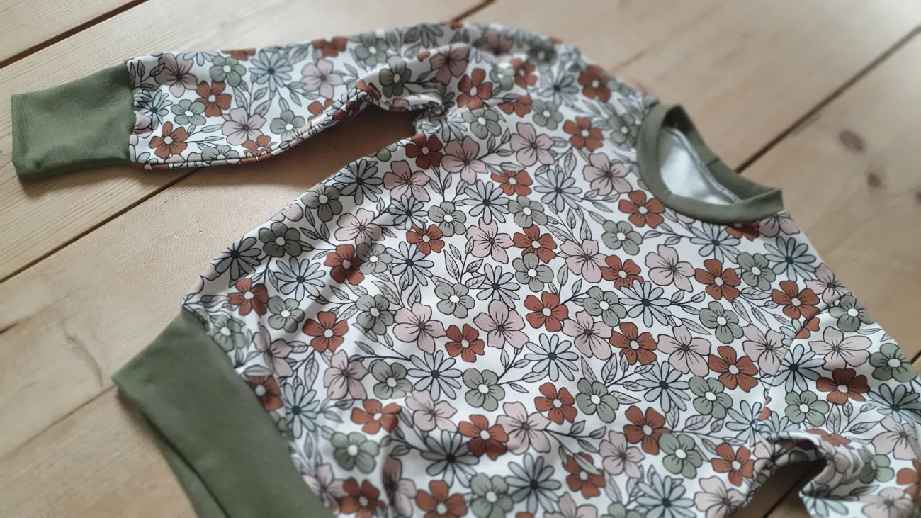 Ein handgefertigter langärmeliger Kinderpullover mit vintage Blumenmuster und dunkelgrünen Bündchen sowie eine cremefarbene Pumphose von ROSA näht, getragen von einem Mädchen auf einer grünen Wiese vor einem Tipi.