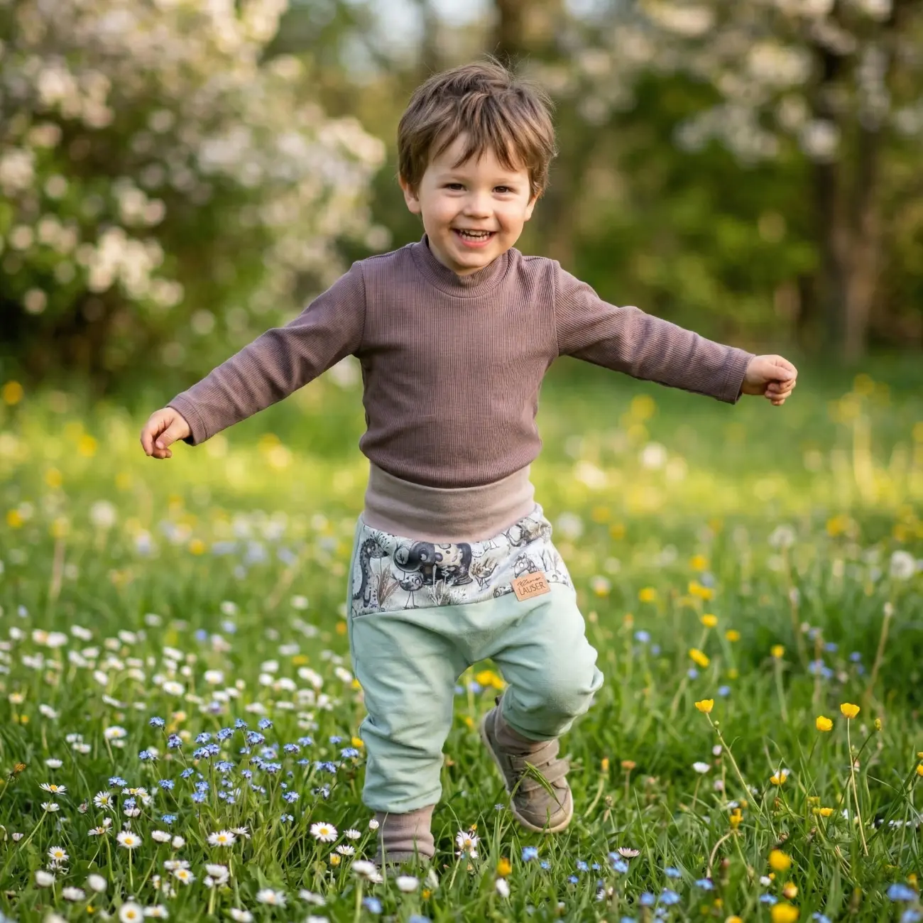 Ein fröhlicher kleiner Junge läuft mit ausgebreiteten Armen über eine Blumenwiese. Er trägt ein Outfit von ROSA näht: ein braunes Langarmshirt aus Rippjersey und eine mitwachsende hellgrüne Pumphose mit breitem beigen Bauchbündchen und einem gemusterten Einsatz im Taschenbereich.
