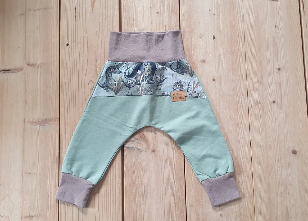 Nahaufnahme einer handgenähten Pumphose von ROSA näht. Die Hose hat hellgrüne Beine, braune Bündchen an Bauch und Knöcheln sowie einen oberen Einsatz aus gemustertem Stoff mit liebevoll gezeichneten Tieren wie Elefanten und kleinen Sauriern.