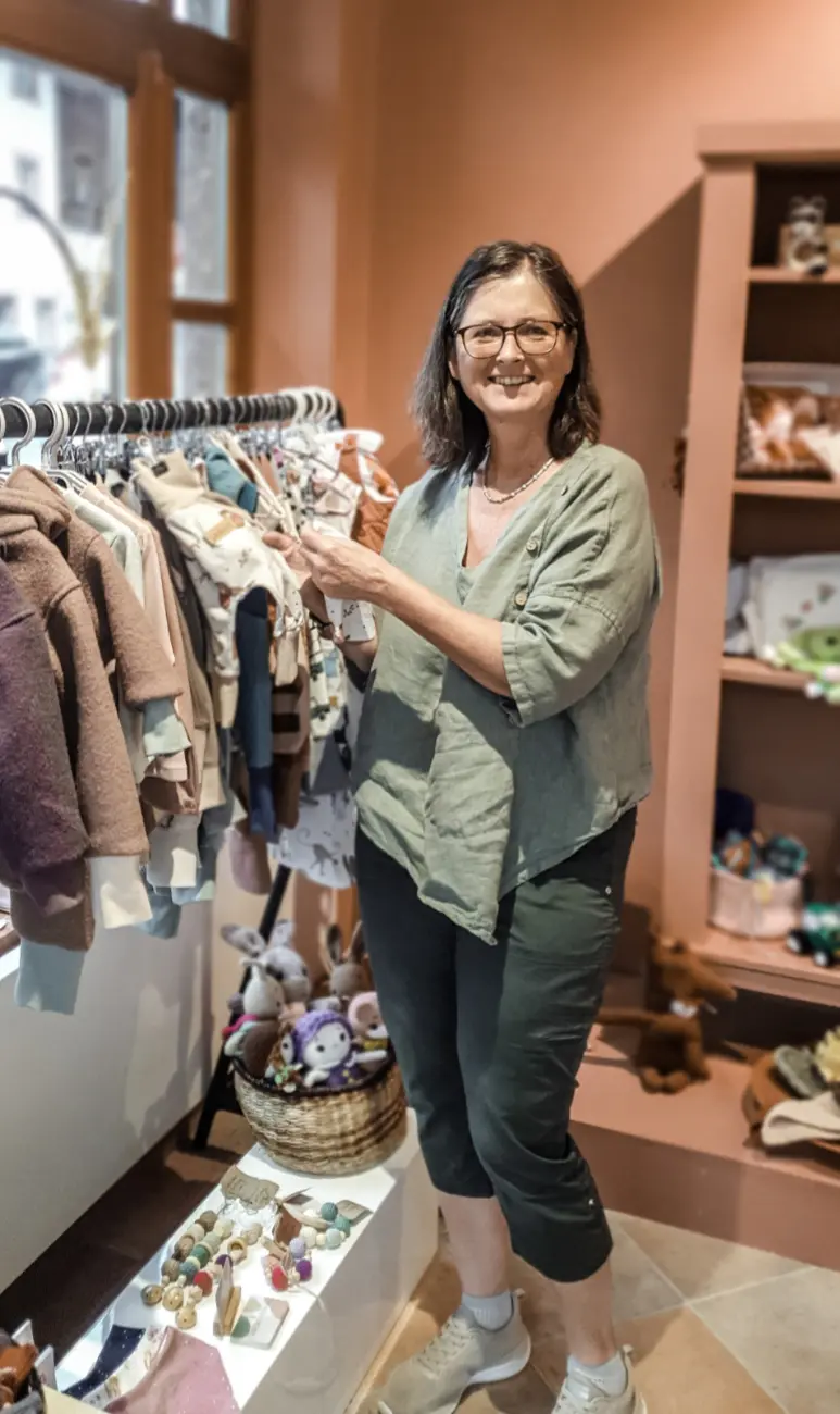 Die Gründerin Roswitha Schwaiger lächelt in ihrem liebevoll eingerichteten Laden für handgemachte Kinderkleidung und gedrechselte Holzprodukte.