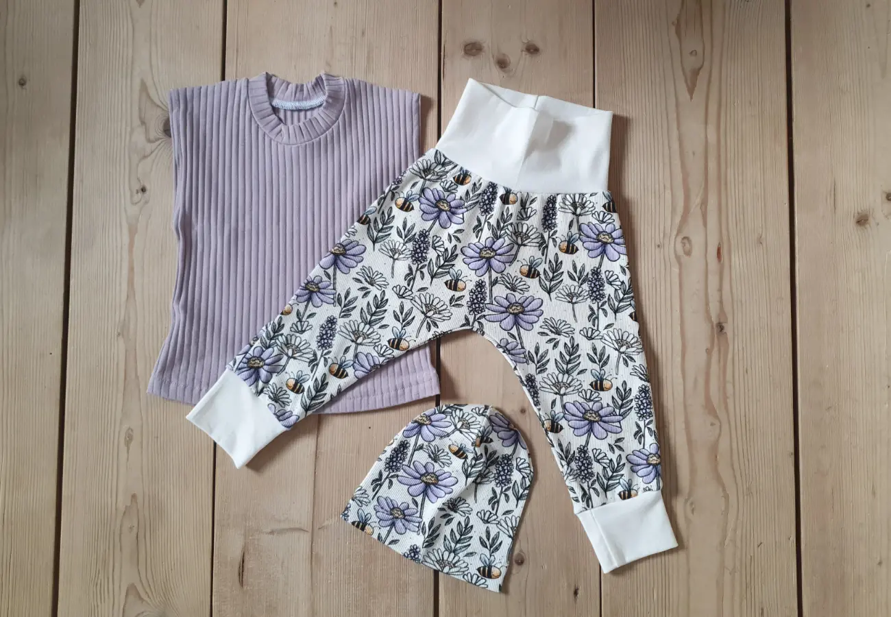 Sommerliches Kinder-Outfit von ROSA näht. Zu sehen ist ein fliederfarbenes, ärmelloses Shirt aus Rippjersey, kombiniert mit einer Pumphose und Mütze. Hosen und Mütze haben helle Bündchen und zeigen ein hübsches Muster mit lilafarbenen Blumen und kleinen Bienen.