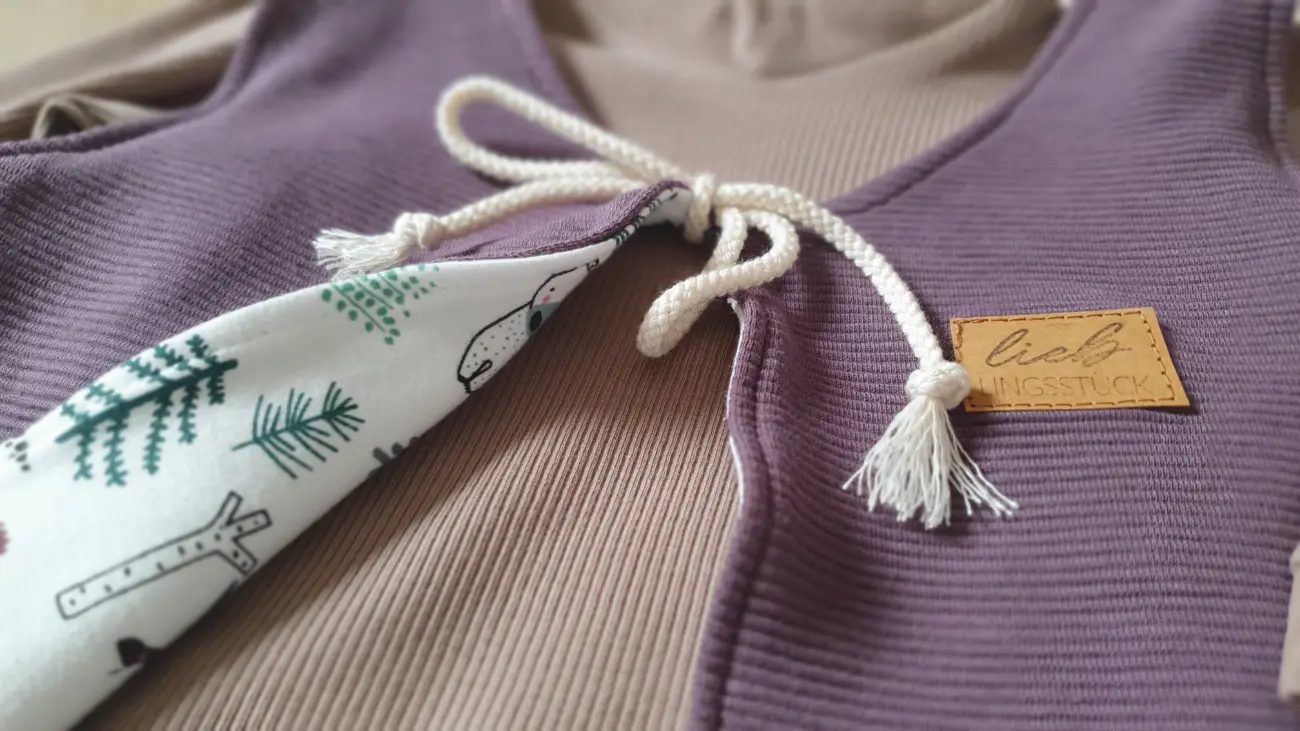 Nahaufnahme einer handgenähten Kinderweste von ROSA näht aus lilafarbenem Stoff mit einer hellen Kordelbindung. Das Innenfutter zeigt ein niedliches Muster mit Waldtieren und Bäumen. Auf der Weste ist ein braunes Kunstleder-Label mit der Aufschrift "Lieblingsstück" angenäht.