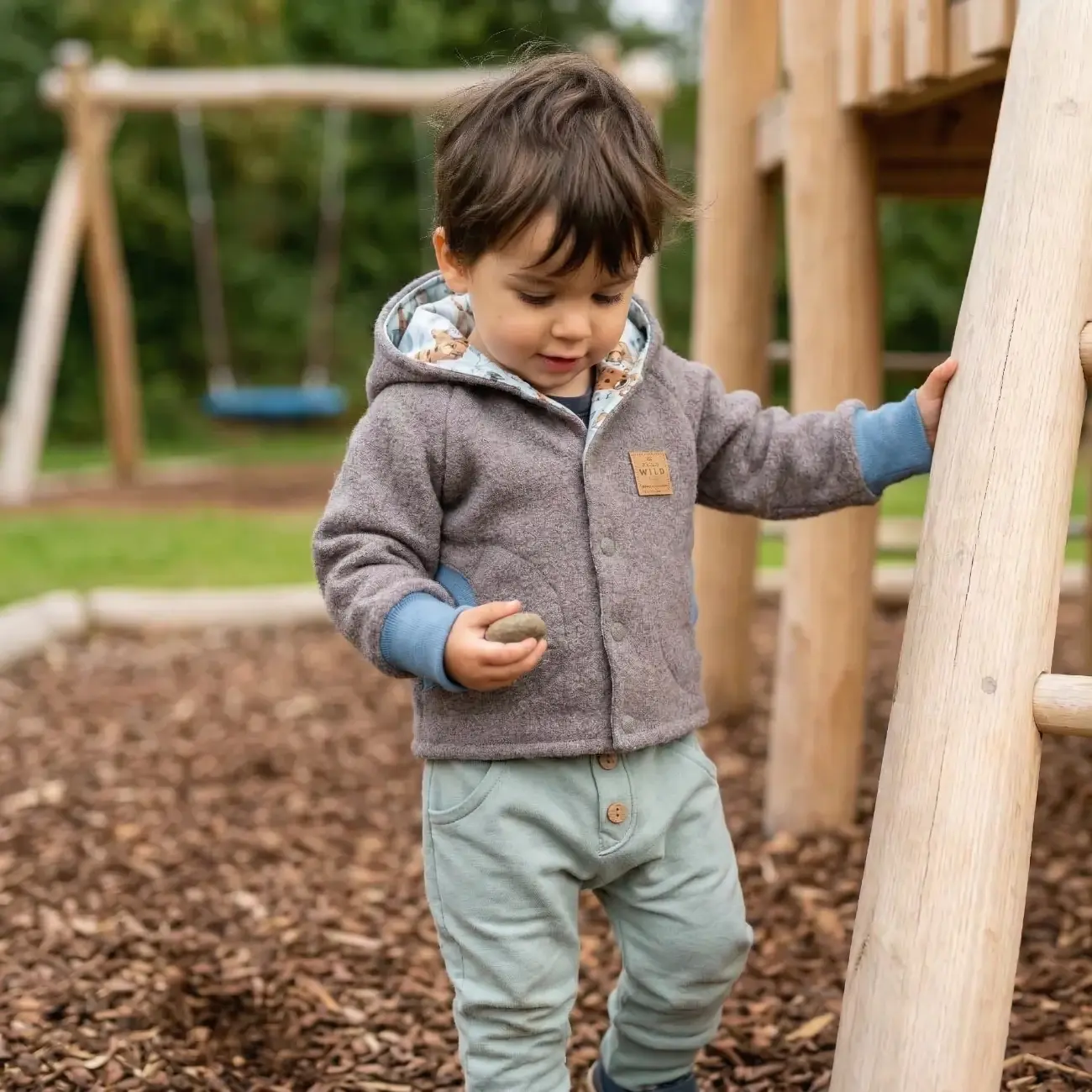 Ein kleiner Junge steht auf einem Holzspielplatz und hält einen Stein. Er trägt eine graue Wollwalkjacke von ROSA näht mit hellblauen Bündchen und einem Innenfutter mit Tiermuster, darunter eine hellgrüne Hose.
