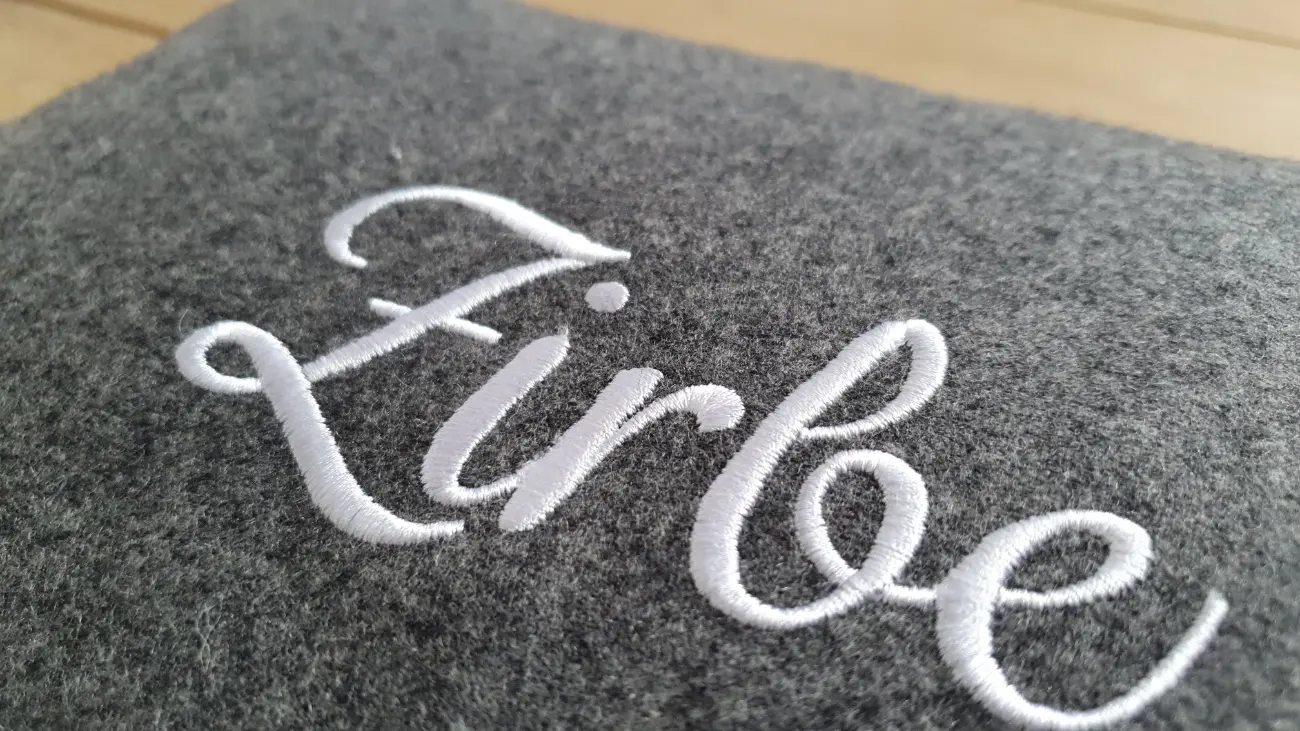 Nahaufnahme der hochwertigen, geschwungenen weißen Stickerei mit dem Schriftzug "Zirbe" auf einem melierten, dunkelgrauen Filzstoff. Ein schönes Detail der handgefertigten Zirbenpolster von ROSA näht.