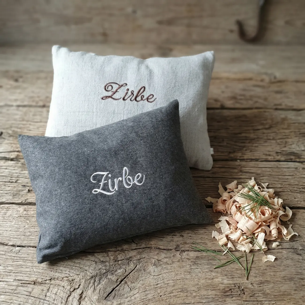 Zwei handgenähte Zirbenpolster von ROSA näht auf einem verwitterten Holztisch. Vorne liegt ein dunkelgraues Kissen aus Filz mit weißer "Zirbe"-Stickerei, dahinter ein naturfarbenes Kissen mit brauner Stickerei. Daneben duftet ein Häufchen frischer Zirbenspäne.